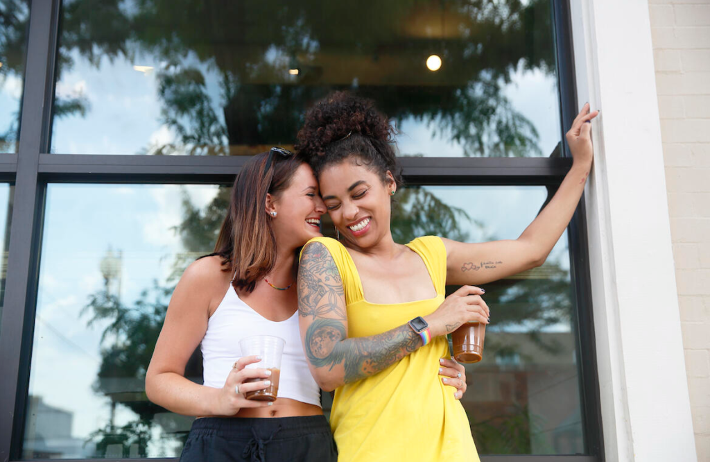 two women hugging on a balcony drinking iced coffees